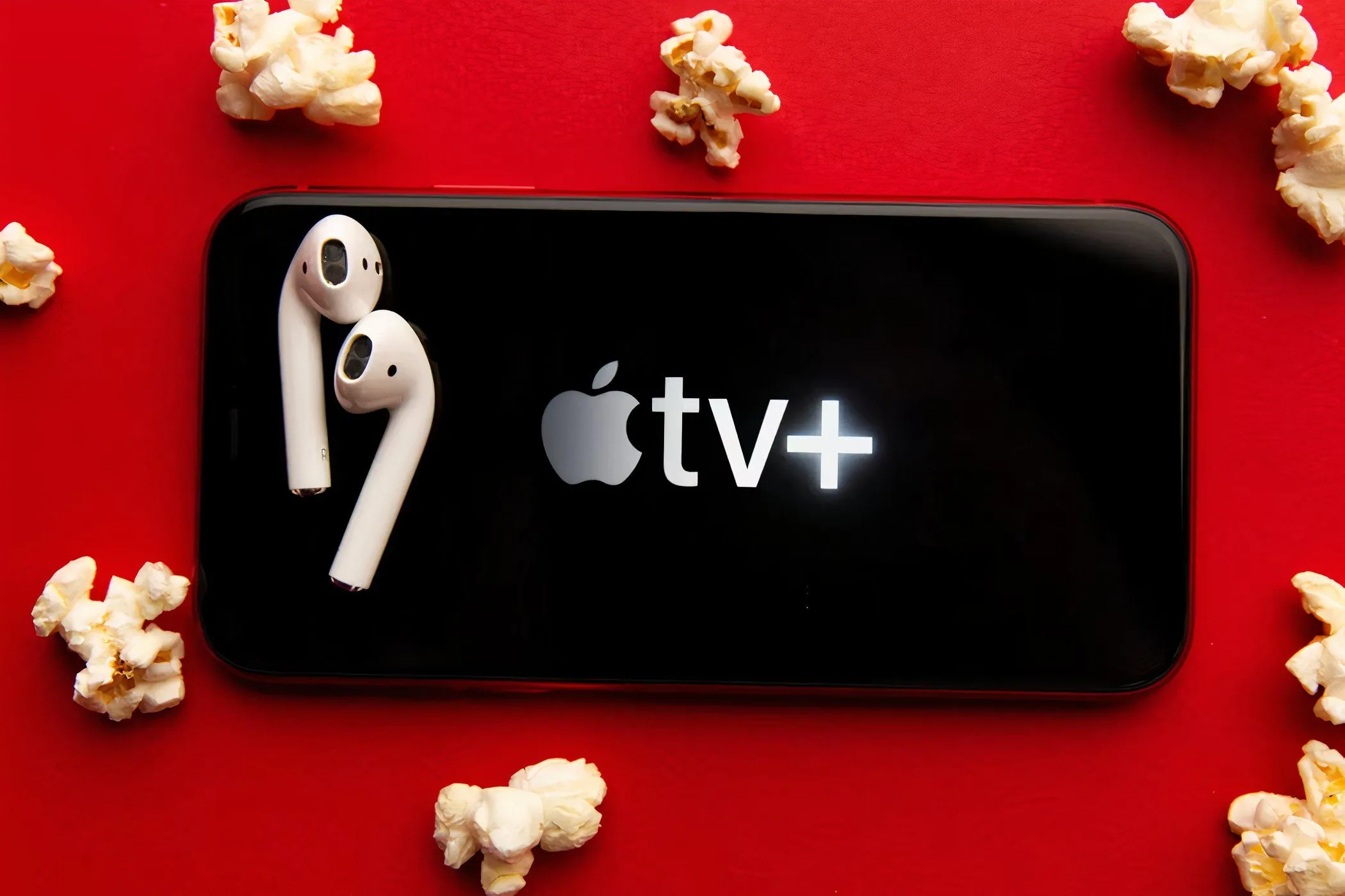 Màn hình điện thoại hiển thị ứng dụng Apple TV+ với logo, xung quanh là bỏng ngô và tai nghe, tượng trưng cho trải nghiệm giải trí tại nhà.